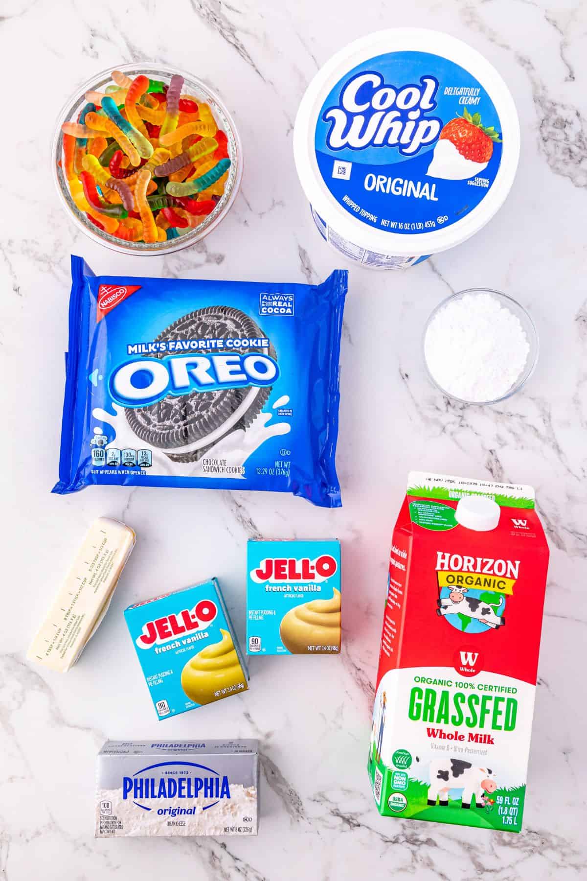 Ingredients on a marble counter gummy worms, Cool Whip, powdered sugar, Oreo cookies, butter, vanilla and chocolate Jell-O pudding, whole milk, and cream cheese.