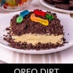 A slice of Oreo Dirt Cake topped with gummy worms is served on a white plate, with a bowl of gummy worms and a stack of Oreos in the background.
