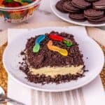An Oreo Dirt Cake with crumbled cookies and gummy worms on top sits on a plate; nearby are a bowl of gummy worms and a plate of chocolate sandwich cookies.