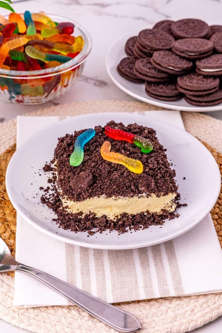 An Oreo Dirt Cake with crumbled cookies and gummy worms on top sits on a plate; nearby are a bowl of gummy worms and a plate of chocolate sandwich cookies.