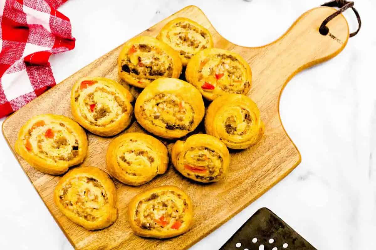 Ten baked pinwheel pastries with a savory sausage, cream cheese, and red pepper filling are arranged on a wooden serving board. A red and white cloth is partially visible in the corner.