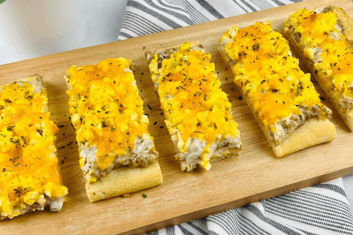 Five rectangular pieces of pizza topped with sausage gravy, scrambled eggs, melted cheddar cheese, and herbs are arranged on a wooden serving board.
