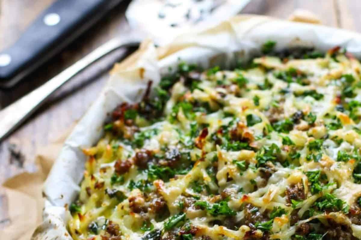 A close-up of a baked crustless topped with melted cheese, chopped kale, sausage, and egg,  with a serving utensil in the background.
