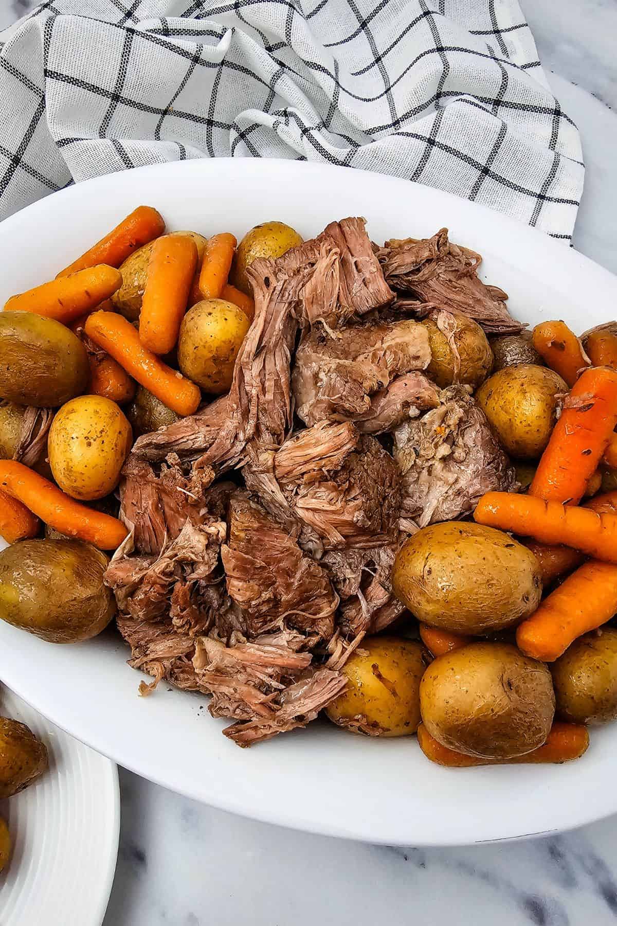A white plate with shredded Slow Cooker Beef Chuck Roast, whole baby potatoes, and cooked carrots set on a marble surface next to a checkered cloth.