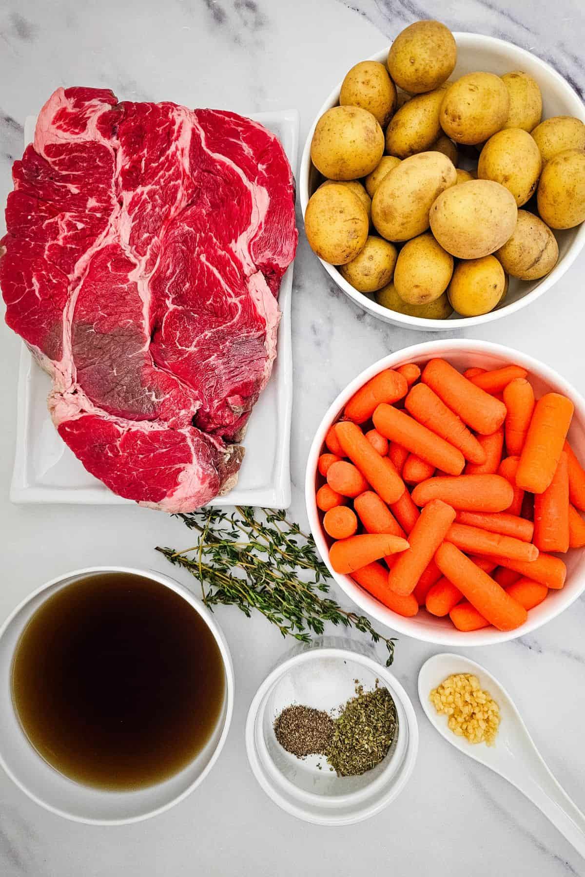 Meat with baby potatoes, carrots, fresh thyme, minced garlic, black pepper, Italian seasoning, and a bowl of broth arranged on a white surface.