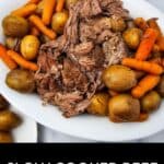 A white platter with Slow Cooker Beef Chuck Roast, whole baby potatoes, and carrots, placed on a marble surface with a checkered cloth in the background.