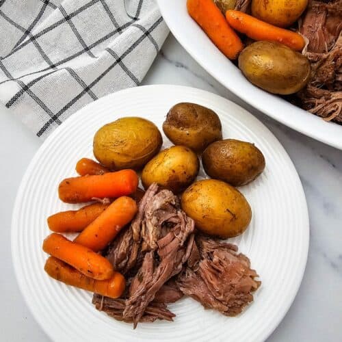 A white plate with cooked baby potatoes, carrots, and Slow Cooker Beef Chuck Roast, next to a serving dish with more of the same food and a checkered cloth.