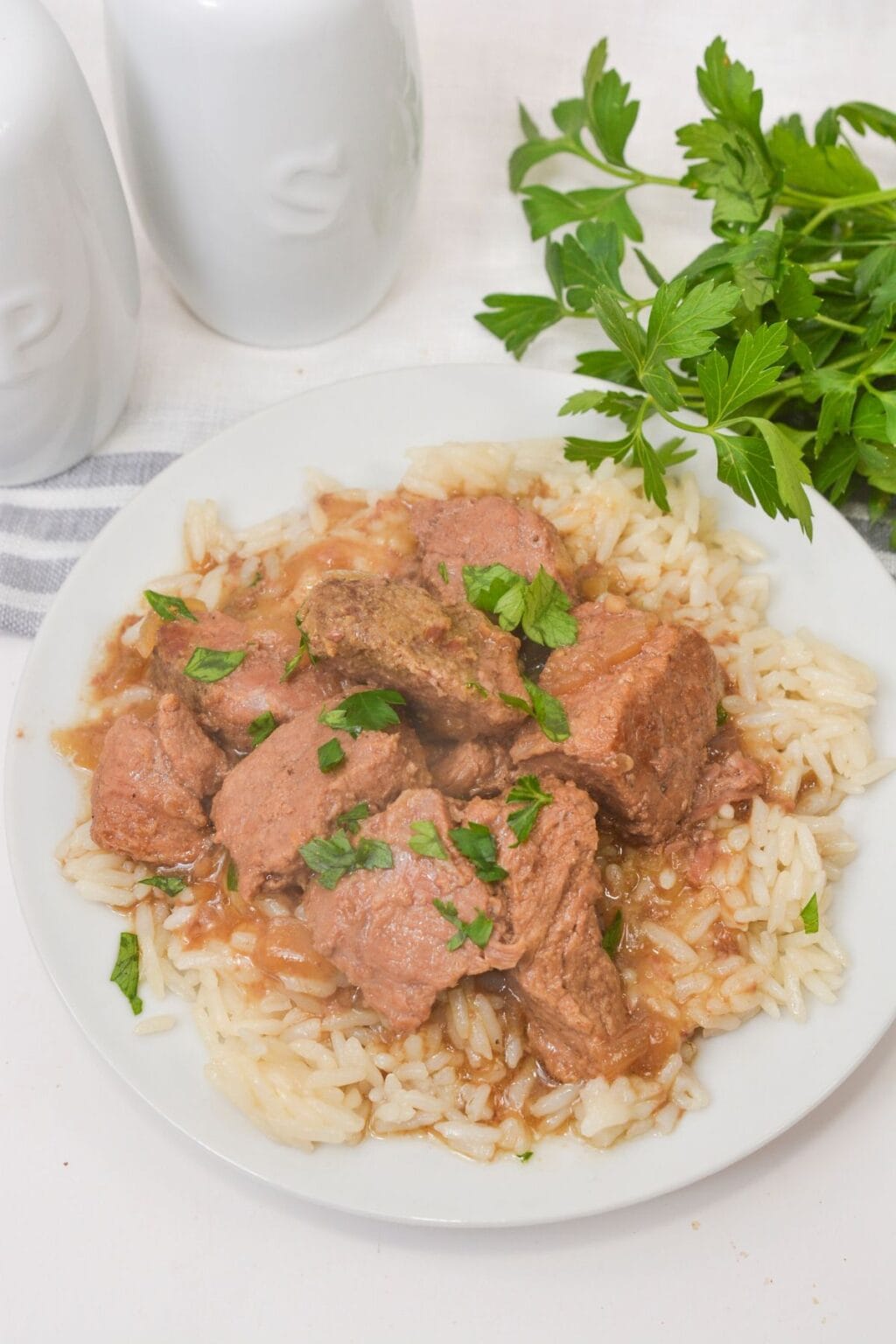 A plate of rice topped with Slow Cooker Beef Tips and Gravy, garnished with chopped parsley, with fresh parsley and salt and pepper shakers in the background.