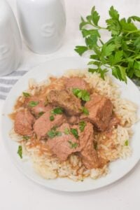 A plate of rice topped with Slow Cooker Beef Tips and Gravy, garnished with chopped parsley, with fresh parsley and salt and pepper shakers in the background.