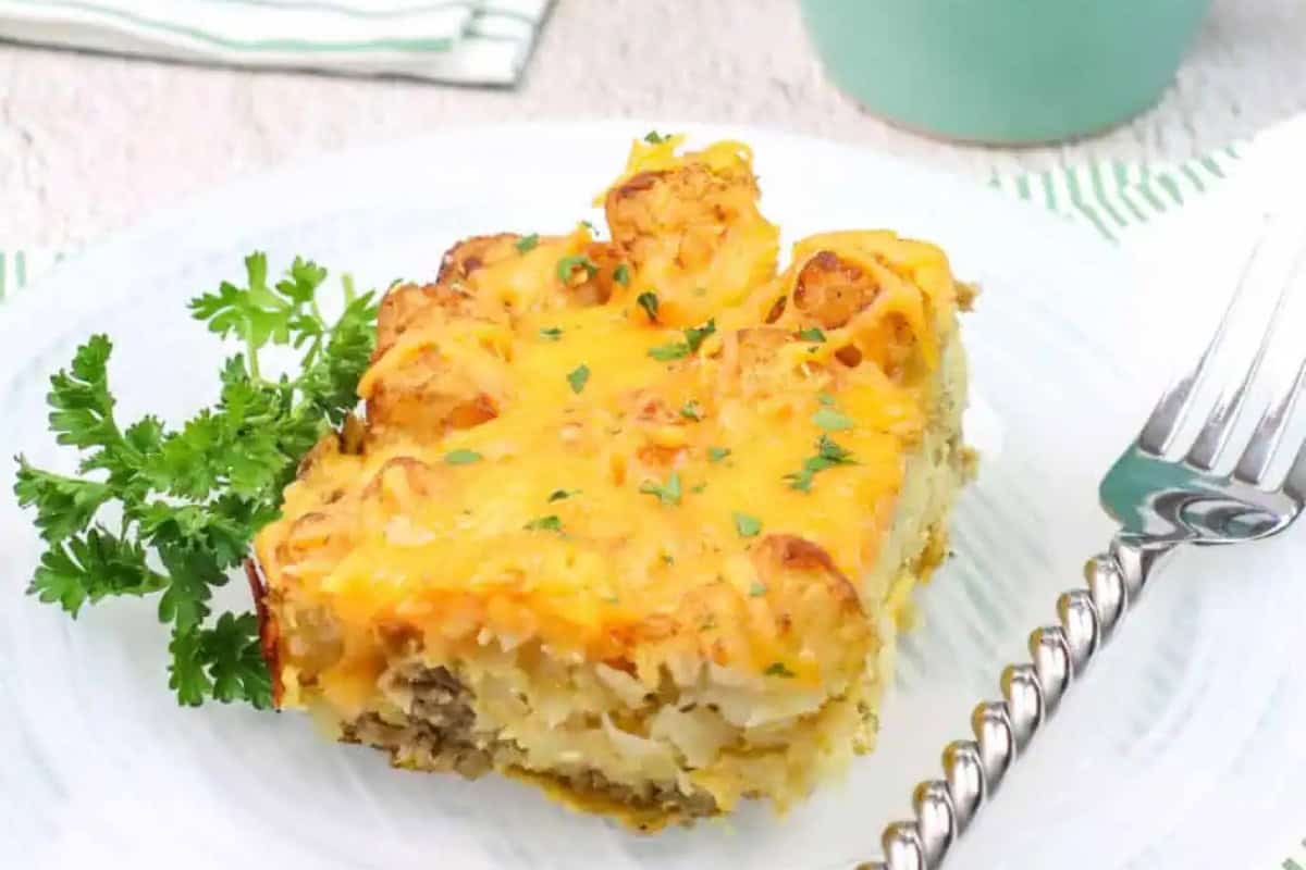 A slice of cheesy tater tot casserole with savory breakfast sausage is garnished with parsley and served on a white plate with a fork.