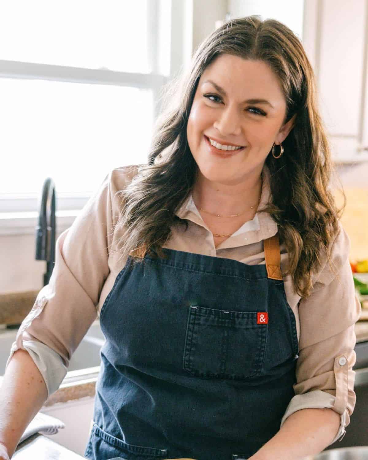 A woman with long brown hair wearing a beige shirt and a dark apron stands in a bright kitchen, smiling at the camera, ready to talk about feeding others and her passion for food on her podcast.
