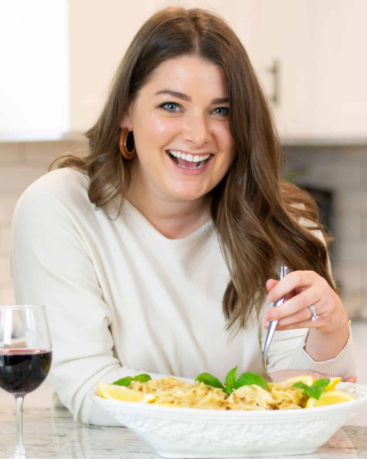 Christina, an inspiration to food bloggers, smiles at the camera with long brown hair, holding a fork near a bowl of pasta. A glass of red wine sits on the table beside her.