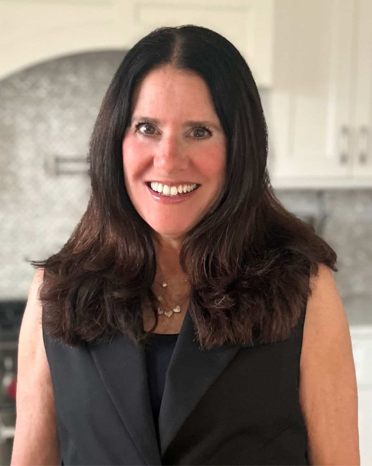 A woman with long dark hair, wearing a black sleeveless top and necklace, smiles at the camera in a kitchen setting, ready to whip up flavorful slow cooker meals.