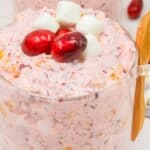 Glass cup filled with pink Cranberry Fluff dessert, topped with two cranberries and three mini marshmallows, with a wooden spoon and some ingredients in the background—perfect as a festive holiday recipe.