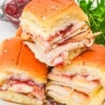 Three Cranberry Turkey Sliders with cheese and cranberry sauce are stacked on a white plate, garnished with parsley, with a bowl of cranberry sauce in the background.