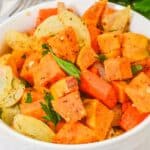 A white bowl filled with Roasted Sweet Potatoes and Carrots, along with onions and parsley, sits on a white surface beside fresh parsley.