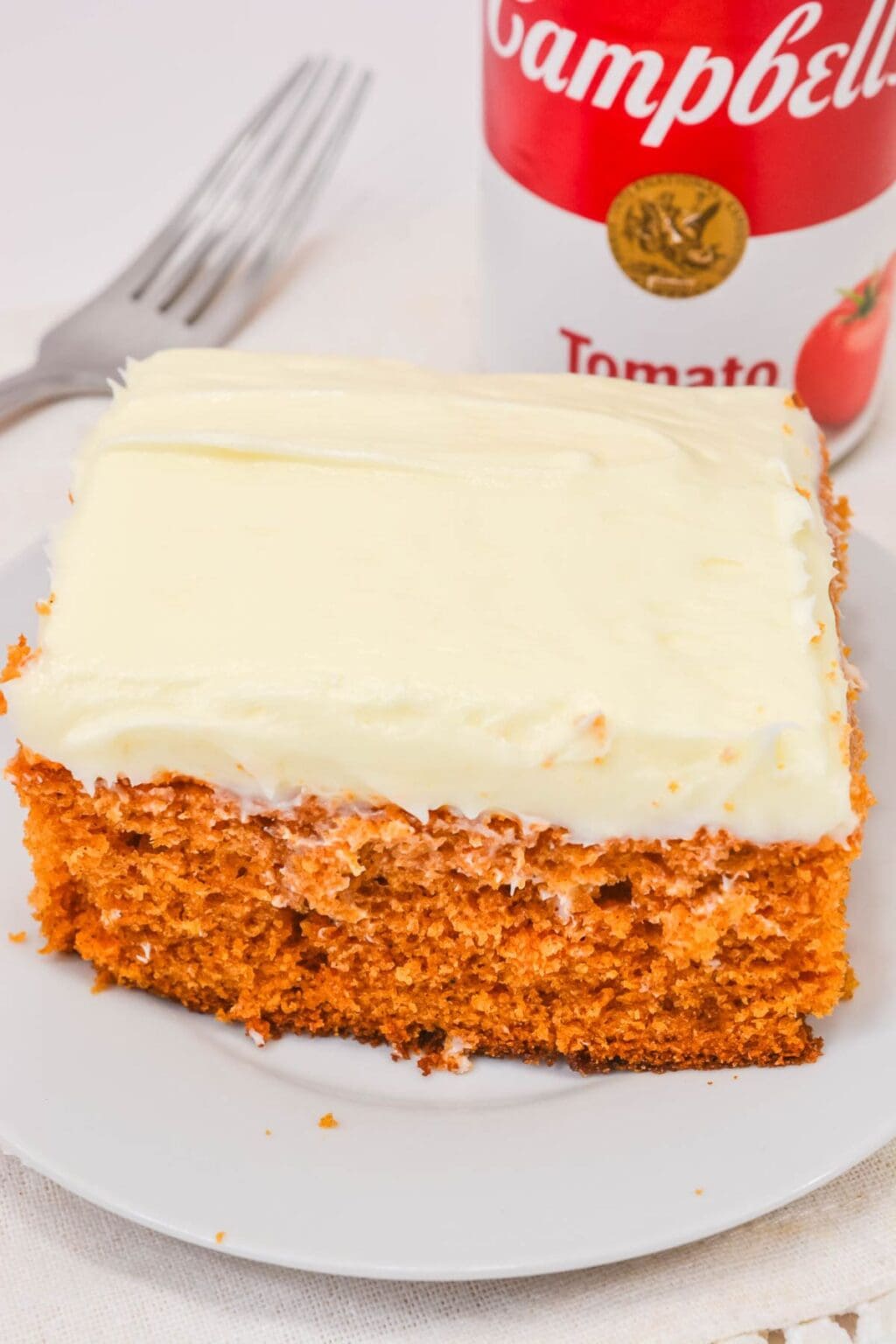 A slice of Vintage Tomato Soup Cake with white frosting sits on a white plate, accompanied by a fork and a can of Campbell's tomato soup in the background.