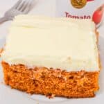 A slice of Vintage Tomato Soup Cake with white frosting sits on a white plate, accompanied by a fork and a can of Campbell's tomato soup in the background.