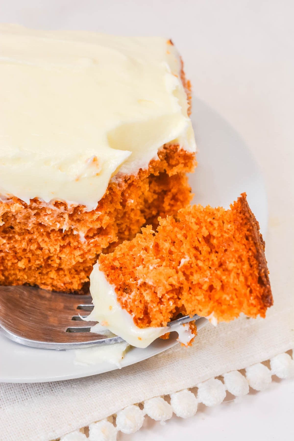 A slice of Vintage Tomato Soup Cake with cream cheese frosting sits on a white plate, a fork holding a bite-sized piece.