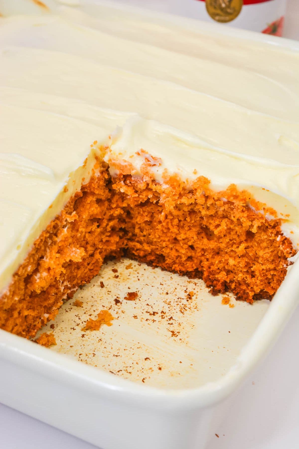 A rectangular pan with white frosting, partially cut to reveal the orange-brown interior, sits in a white baking dish.