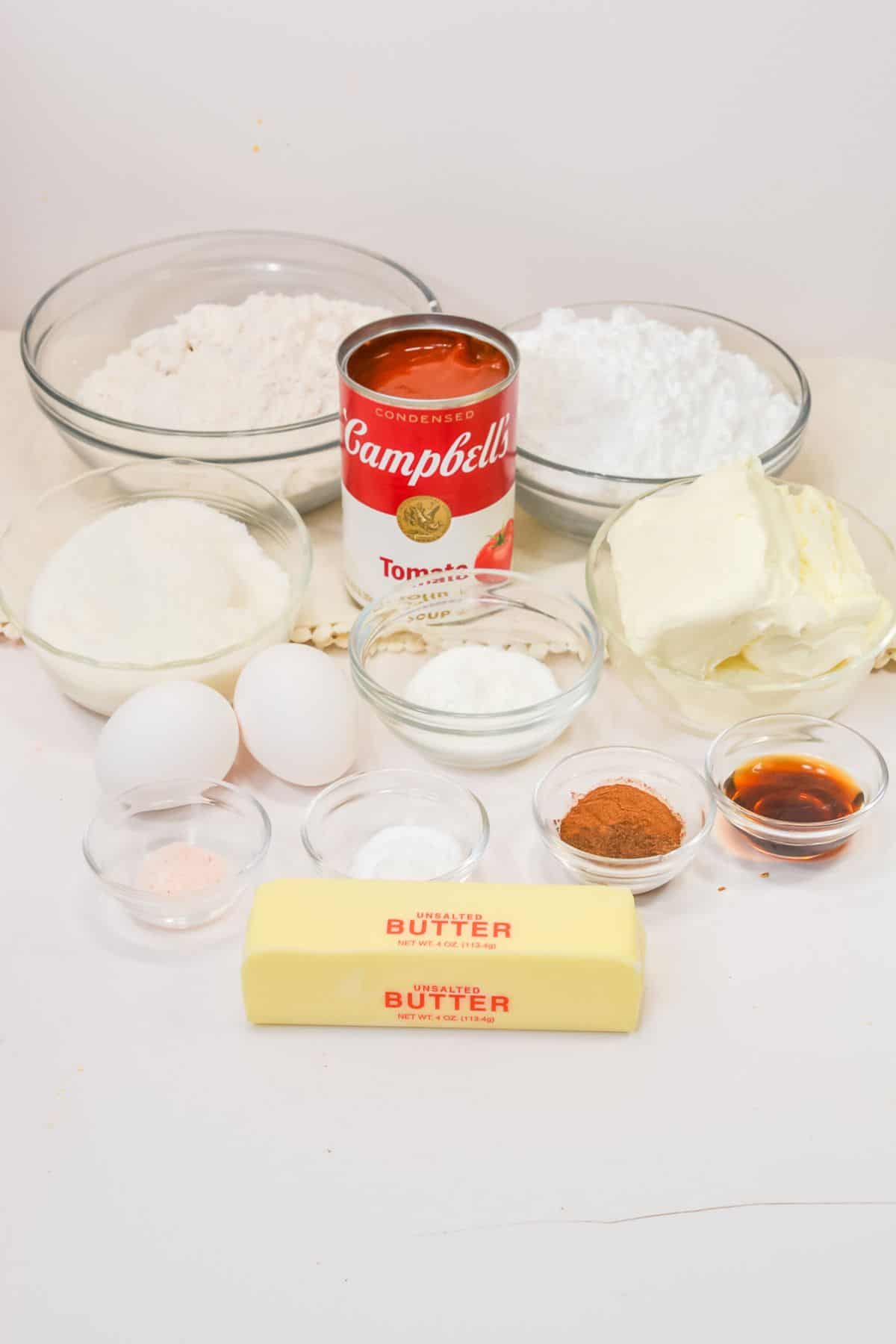 Assorted baking ingredients for Vintage Tomato Soup Cake—including a can of tomato soup, stick of butter, eggs, bowls of flour, sugar, baking soda, spices, vanilla extract, and cream cheese—are arranged on a white surface.