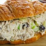 A croissant sandwich filled with Cranberry Chicken Salad—featuring tender chicken, celery, and dried cranberries—rests on a wooden surface beside a colorful kitchen towel.