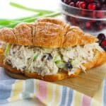 A croissant sandwich filled with cranberry chicken salad, placed on a wooden board with a bowl of cranberries and green onions in the background.