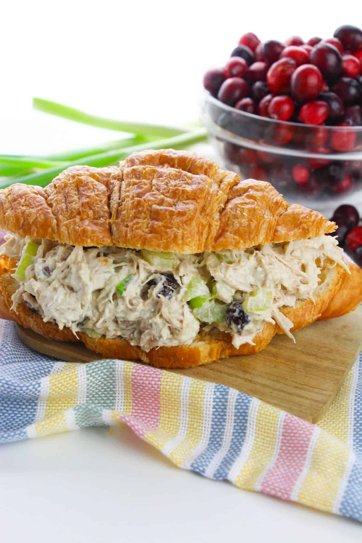 A croissant sandwich filled with cranberry chicken salad, placed on a wooden board with a bowl of cranberries and green onions in the background.