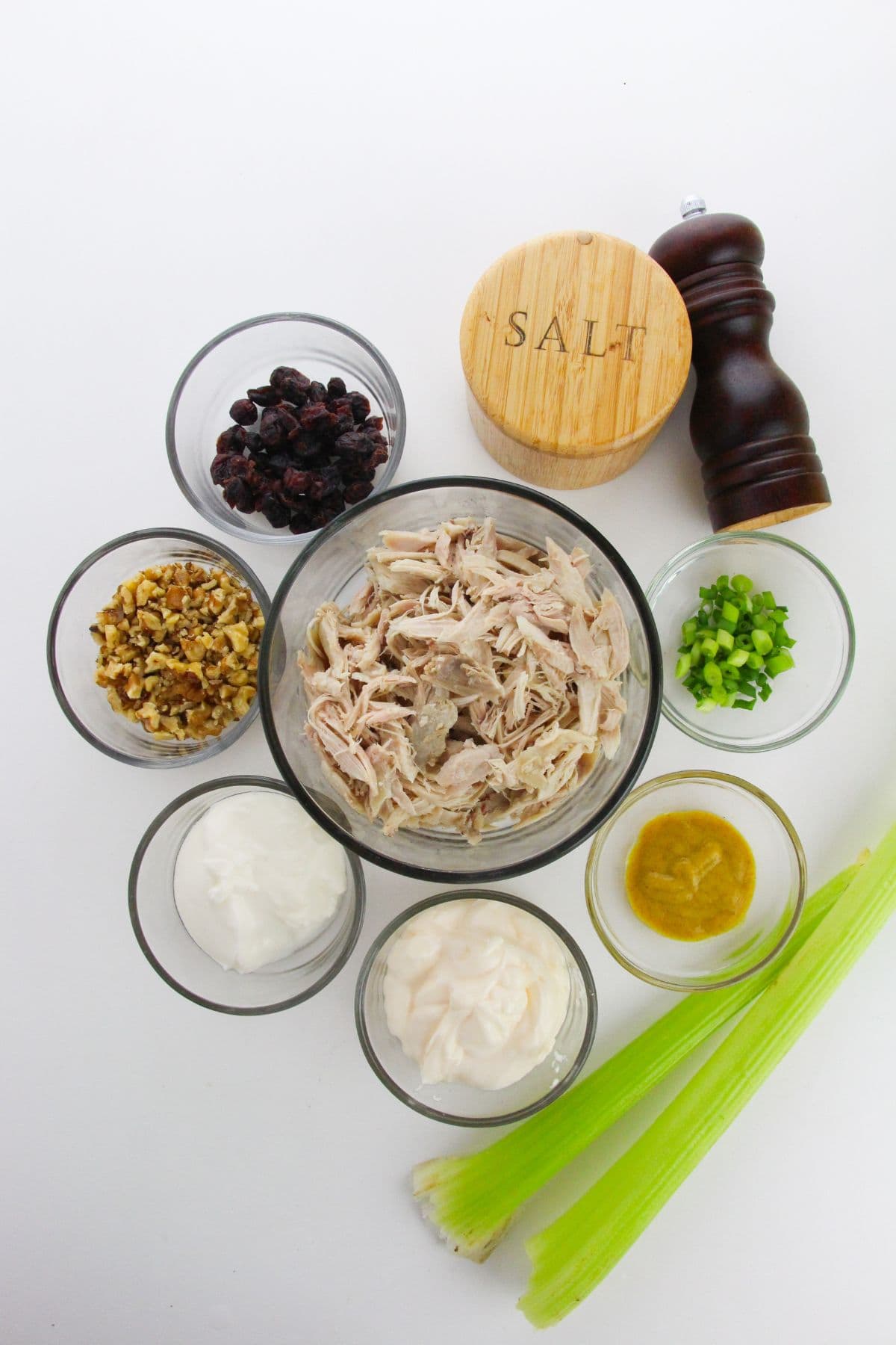 Ingredients in bowls: Meat, mayonnaise, yogurt, mustard, chopped green onions, dried cranberries, walnuts, salt, pepper, and celery sticks on a white background.