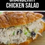 A croissant sandwich filled with Cranberry Chicken Salad sits on a wooden board with a colorful cloth underneath.