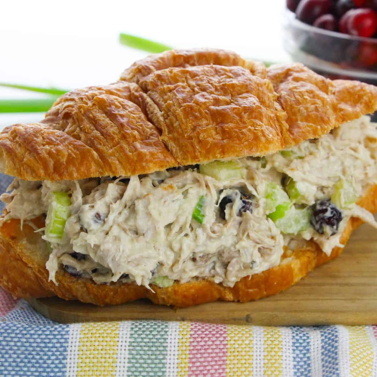 A Cranberry Chicken Salad croissant sandwich, filled with chicken salad, celery, and dried cranberries, sits on a wooden board with a colorful napkin.