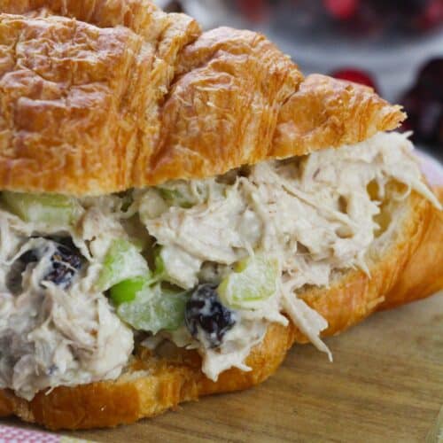 A croissant sandwich filled with Cranberry Chicken Salad—featuring celery and dried cranberries—rests on a wooden surface near a striped cloth.