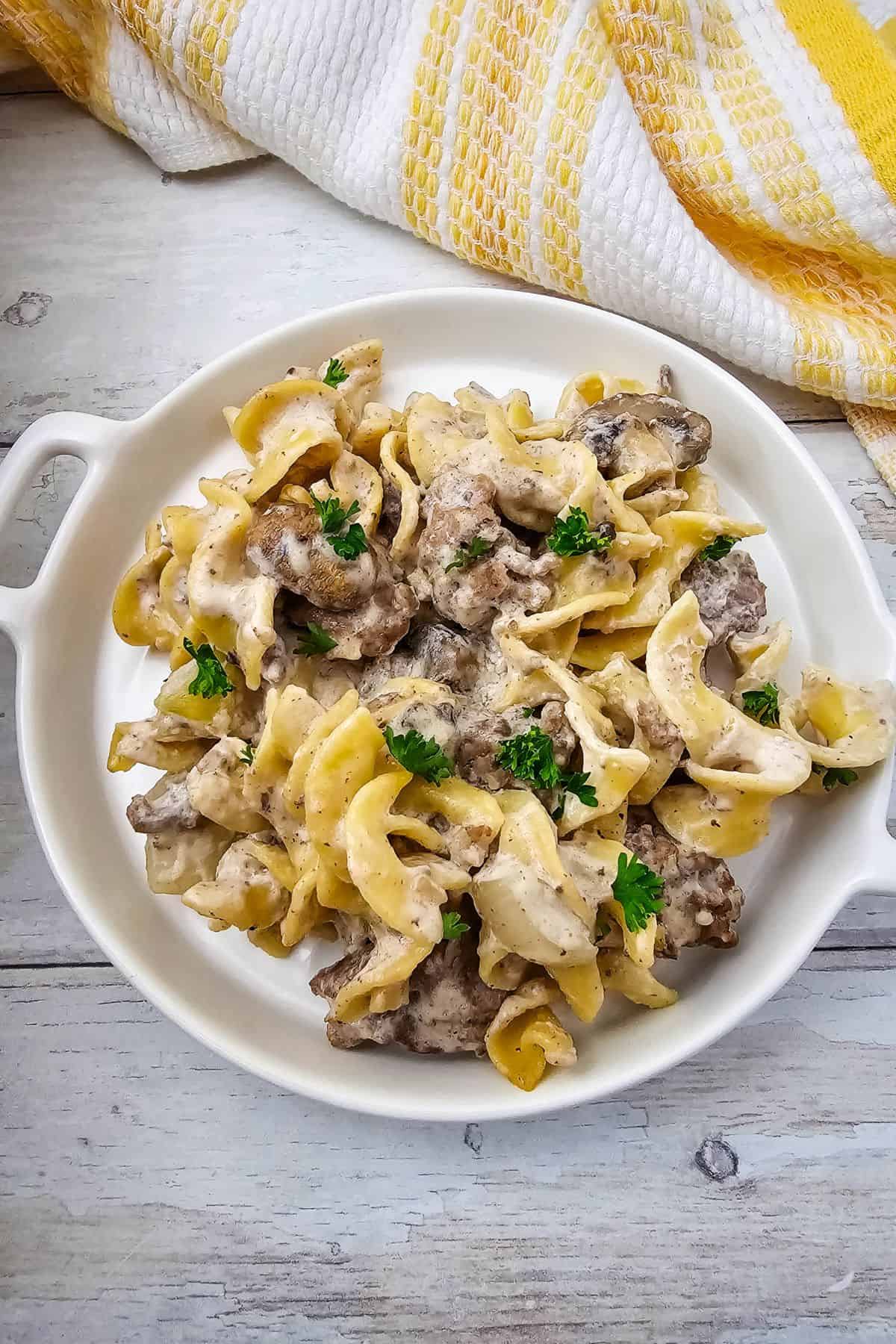 A white plate of Easy Ground Beef Stroganoff with egg noodles, creamy sauce, and parsley garnish sits on a light wooden table next to a yellow and white striped towel.