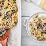 A white baking dish filled with creamy Easy Ground Beef Stroganoff noodle casserole is partially served onto a white plate, with a wooden spoon and a yellow-striped towel nearby.