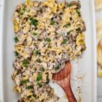 A casserole dish filled with creamy Easy Ground Beef Stroganoff and noodles, garnished with parsley, with a wooden serving spoon partially scooping a portion.