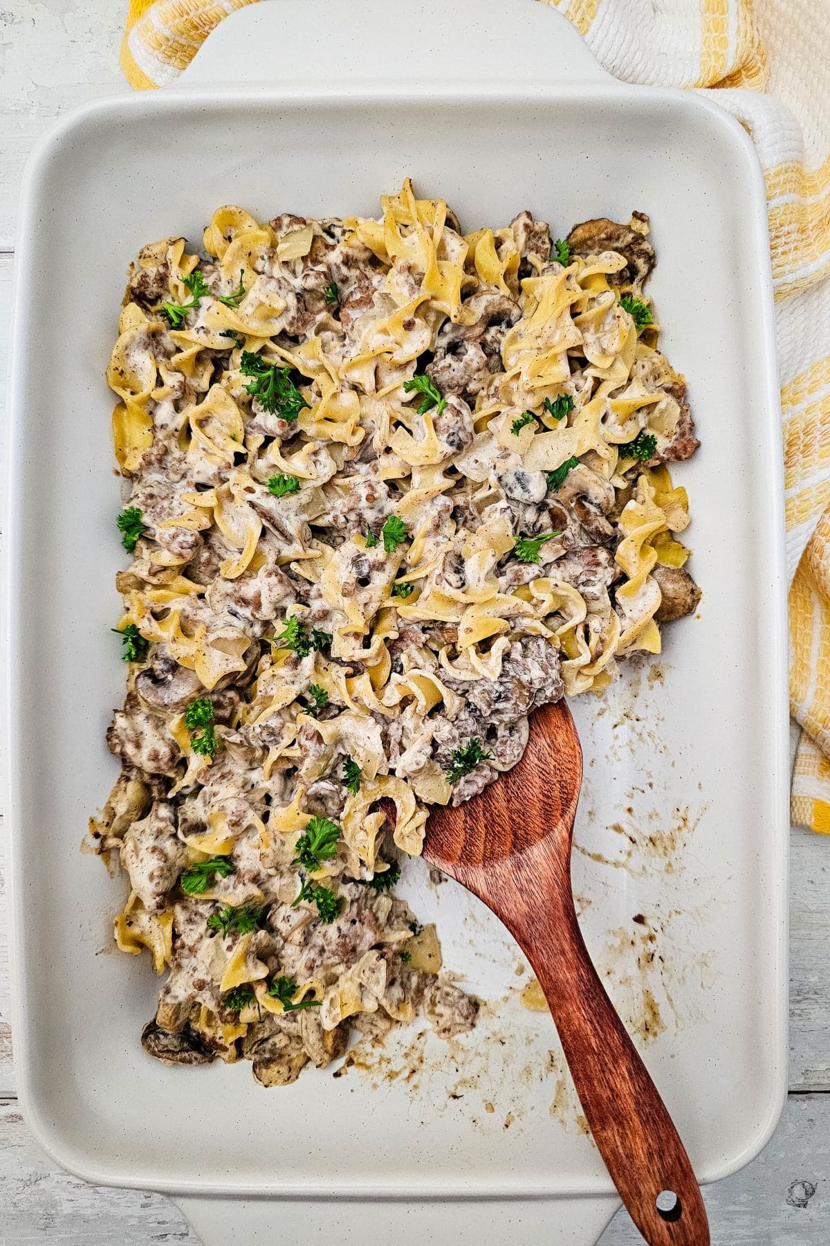 A casserole dish filled with creamy Easy Ground Beef Stroganoff and noodles, garnished with parsley, with a wooden serving spoon partially scooping a portion.