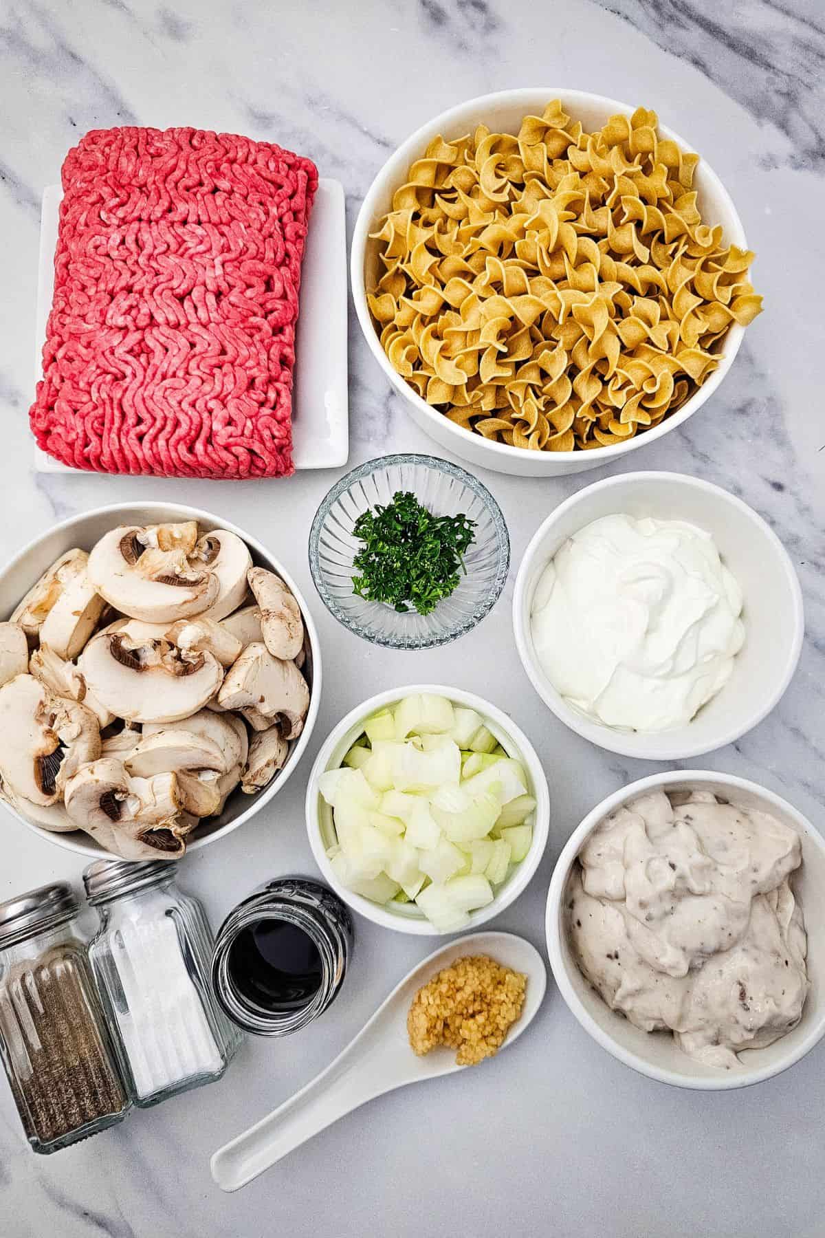 Ingredients for an Easy Ground Beef Stroganoff are arranged on a countertop, including ground beef, egg noodles, mushrooms, onions, garlic, herbs, sour cream, soup, Worcestershire sauce, salt, and pepper.