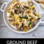 A white dish filled with Easy Ground Beef Stroganoff casserole, garnished with parsley, sits on a light wooden surface next to a yellow and white towel.