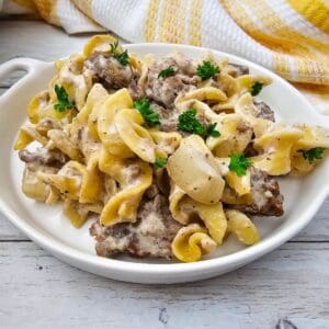 A white plate with Easy Ground Beef Stroganoff, creamy egg noodles, mushrooms, and parsley garnish sits on a light wooden surface with a yellow and white cloth in the background.