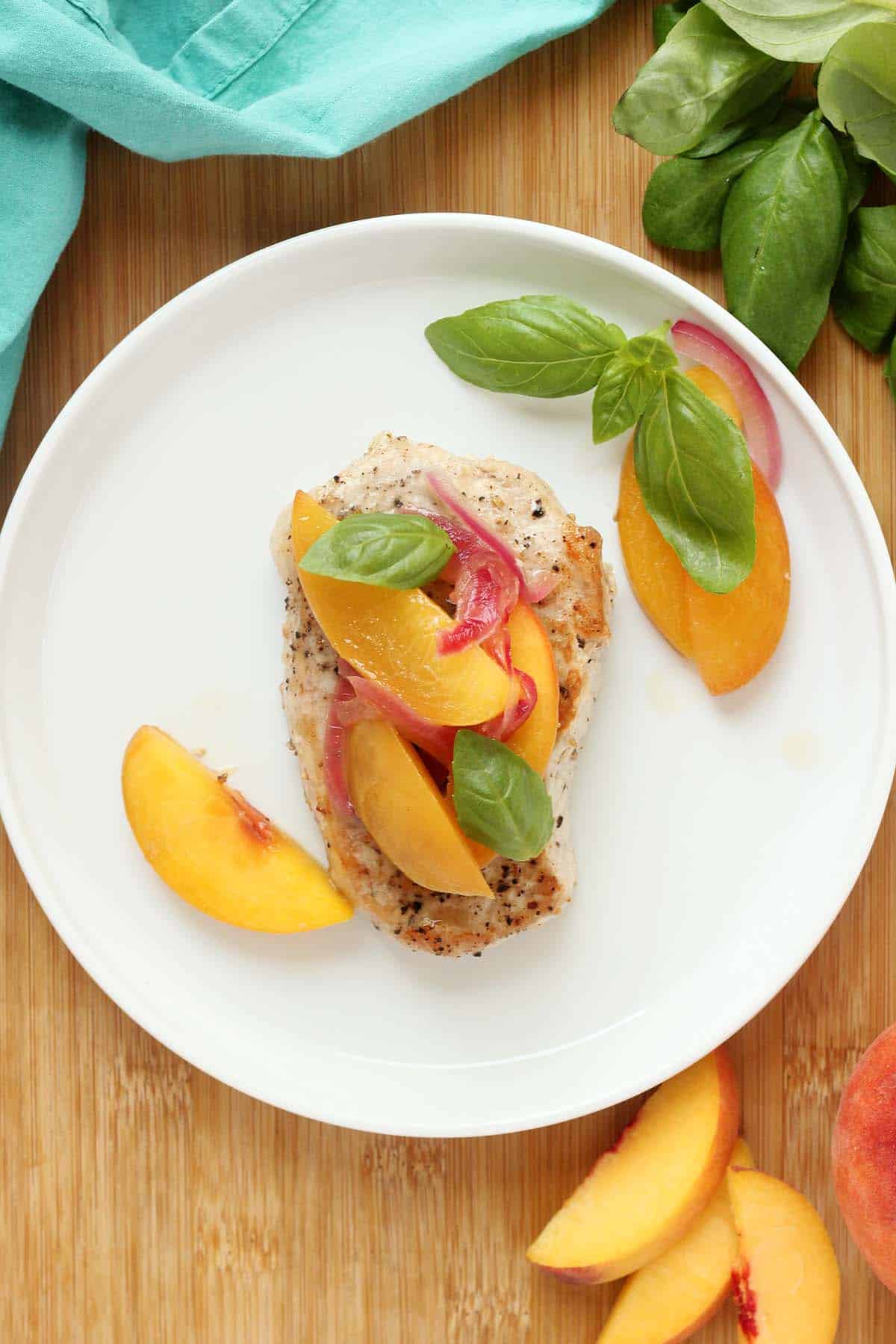 Grilled chicken breast topped with peach slices, red onion, and basil leaves is beautifully arranged on a white plate, with extra peaches and basil nearby on a wooden surface. While this dish features chicken, you can easily adapt it using delicious pork chops for a twist inspired by your favorite pork chop recipes.