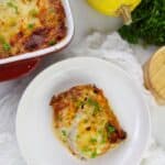 A serving with melted cheese and herbs on a white plate, next to a casserole dish, spaghetti squash, and fresh parsley.