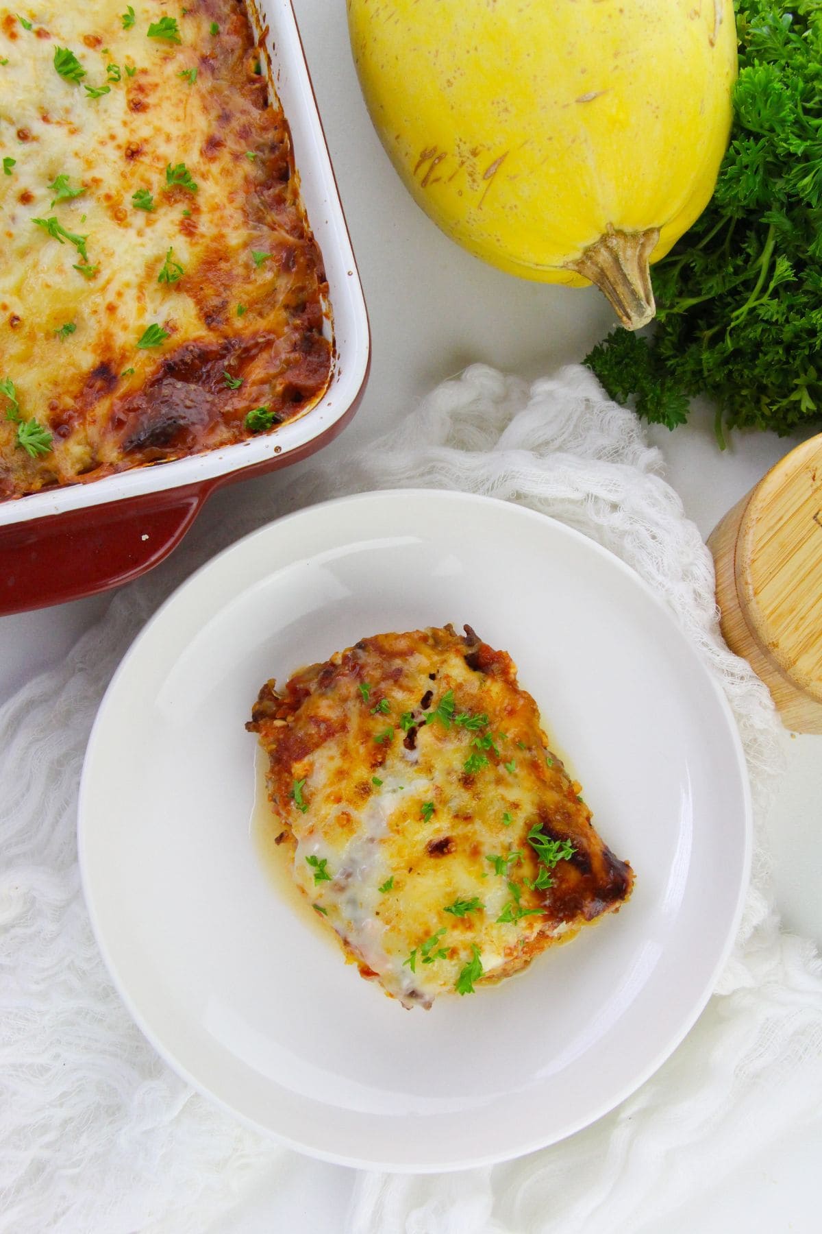 A serving  with melted cheese and herbs on a white plate, next to a casserole dish, spaghetti squash, and fresh parsley.