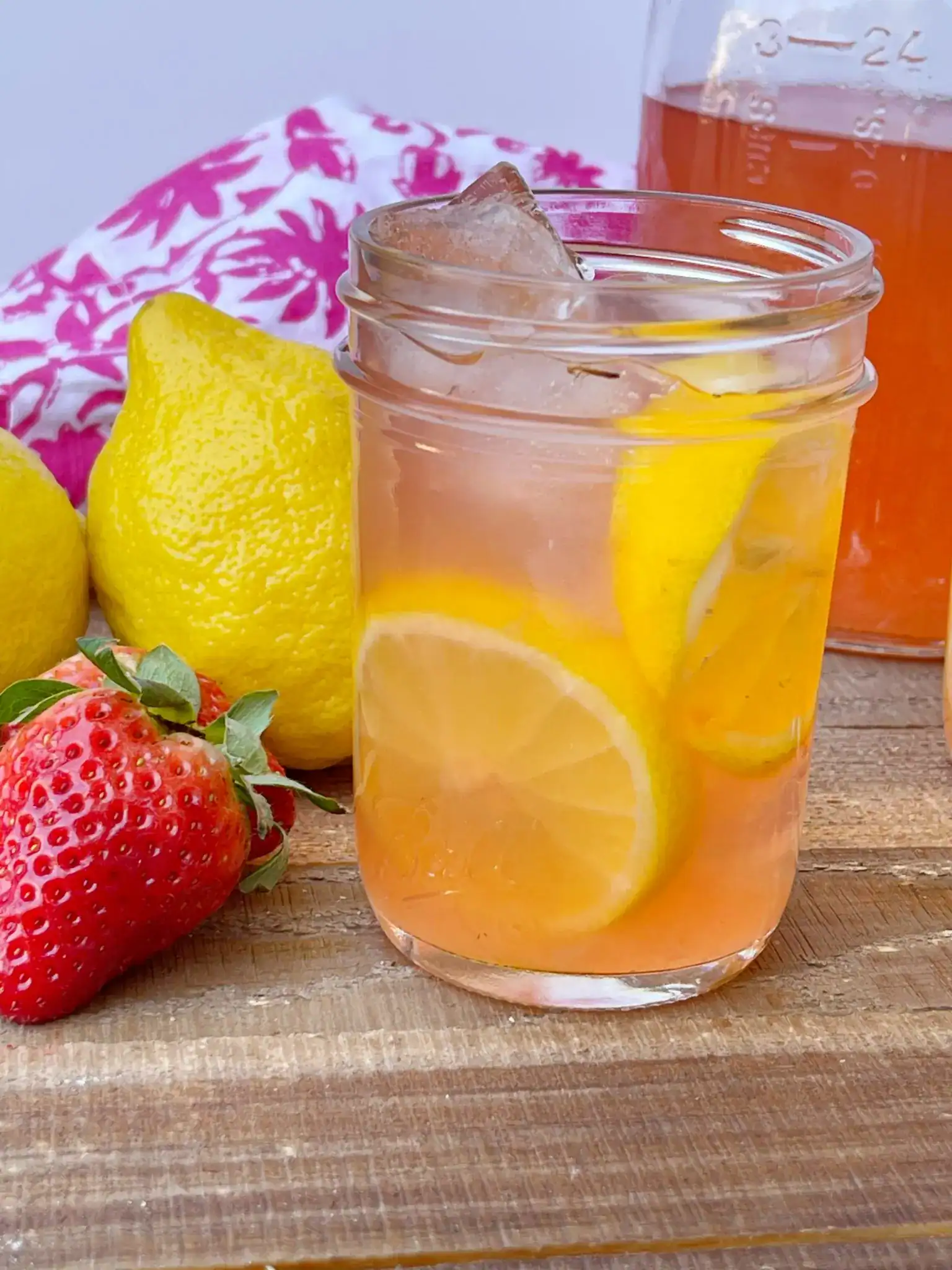 A glass of iced lemonade with lemon slices on a wooden surface, surrounded by whole lemons, a strawberry, and a floral cloth in the background—perfect inspiration for creating your own flavored moonshine recipes.