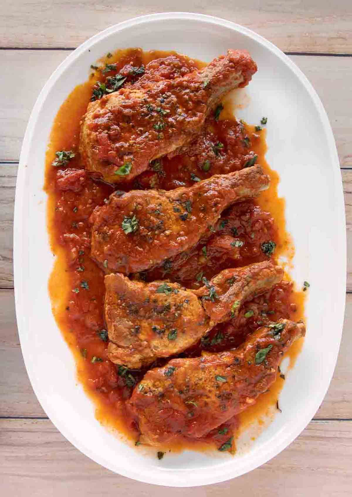 Four pieces of braised chicken in tomato sauce are arranged on a white oval plate, garnished with chopped herbs—an inviting presentation reminiscent of delicious pork chops featured in easy pork recipes.