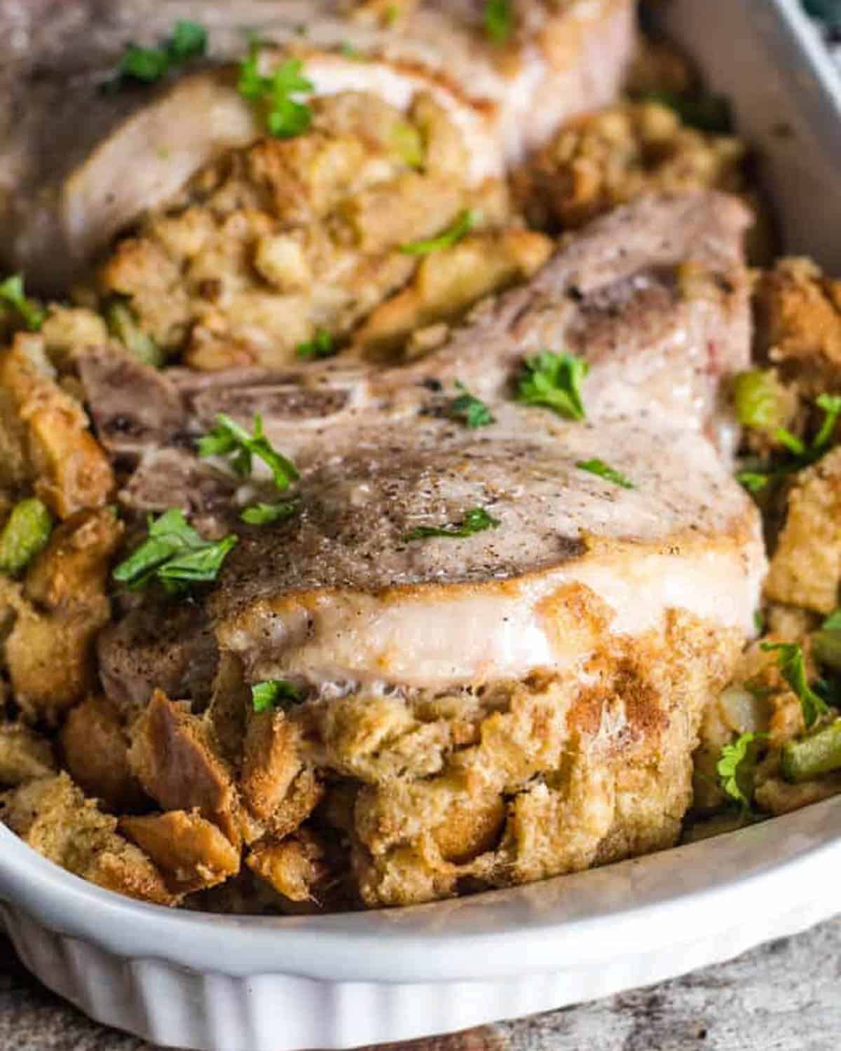 Baked pork chops with stuffing and chopped herbs in a white casserole dish make for one of the most delicious pork chop recipes you’ll ever try.