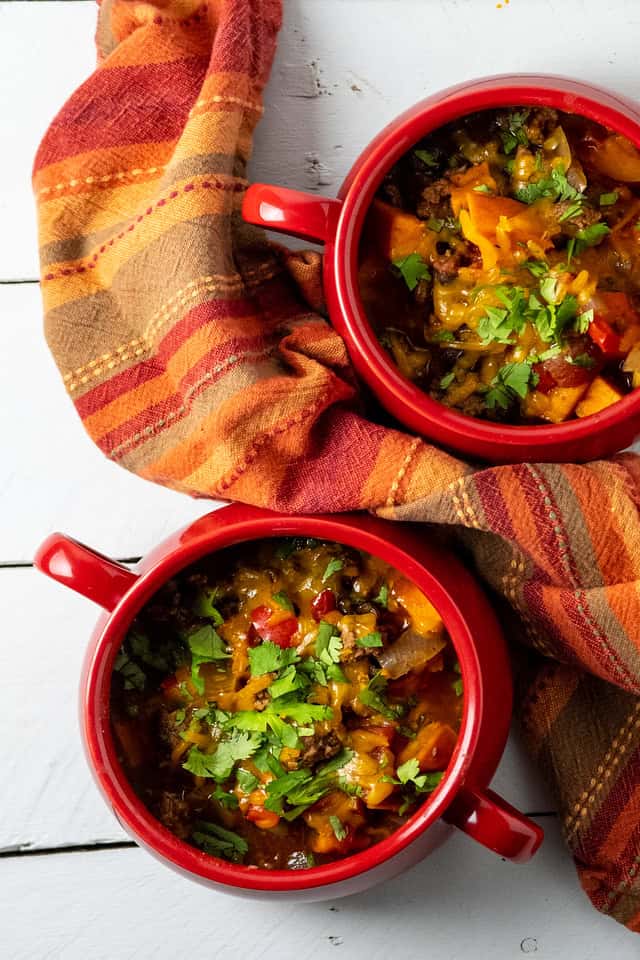 Two red bowls filled with chunky vegetable stew—a must try for fans of chili recipes—topped with chopped herbs, placed on a white wooden surface with a red and orange striped cloth.