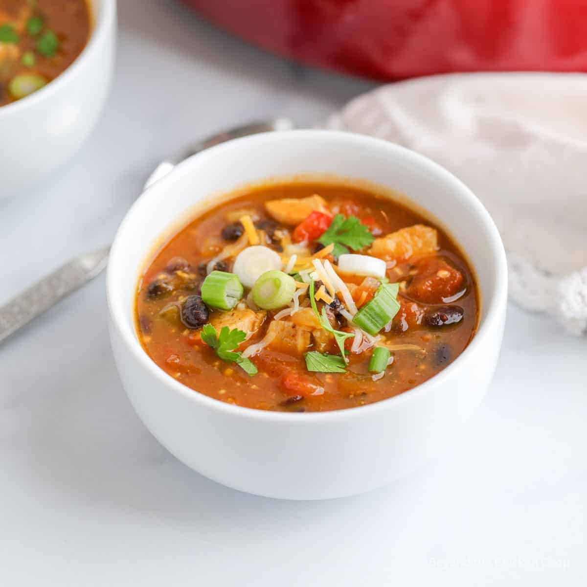 A white bowl filled with chicken chili, black beans, tomatoes, and topped with shredded cheese, green onions, and parsley—this must try chili is a standout among popular chili dishes.