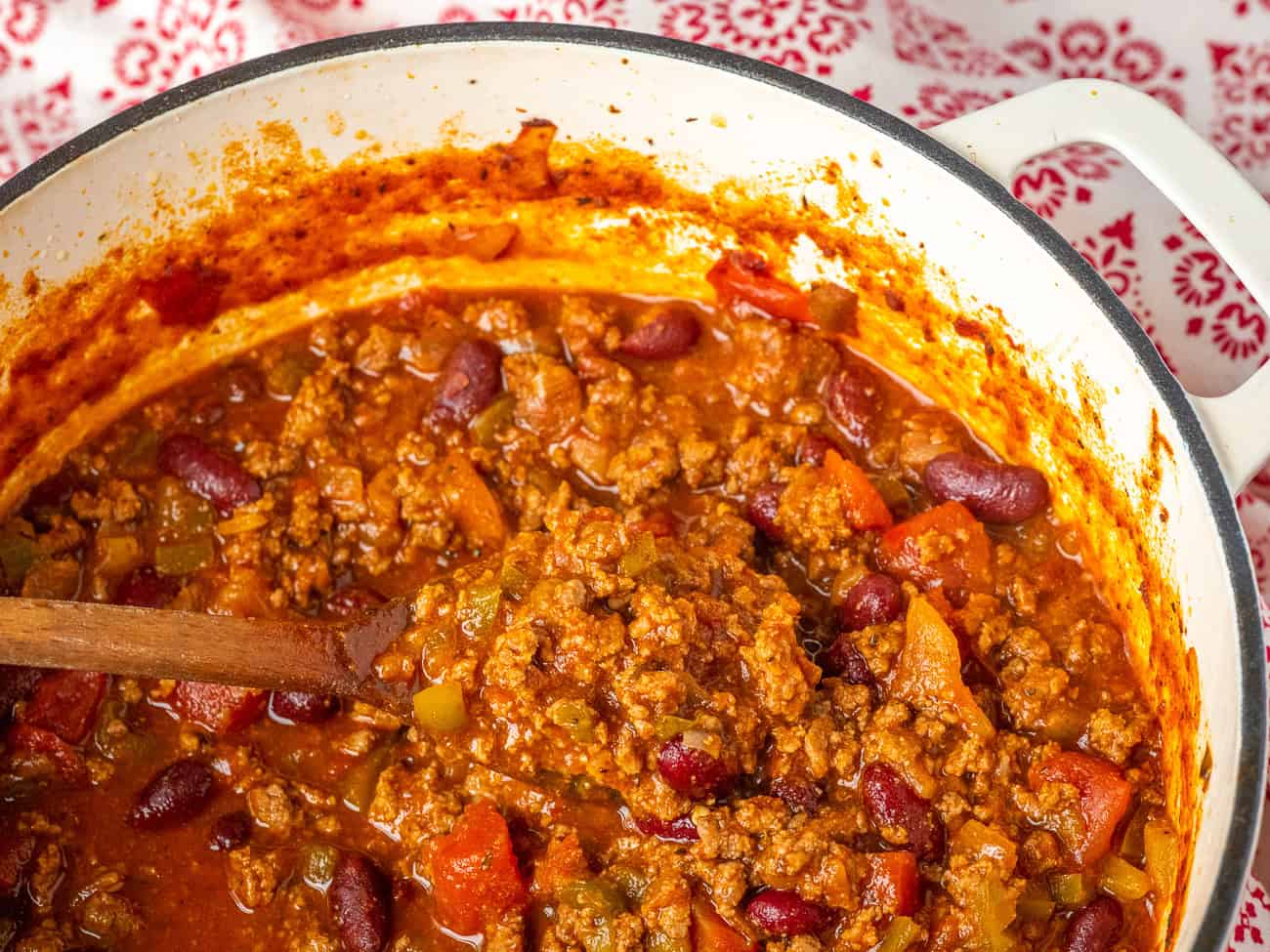 A pot of popular chili with ground meat, kidney beans, peppers, and tomatoes is being stirred with a wooden spoon—one of the must try chili recipes for any comfort food lover.