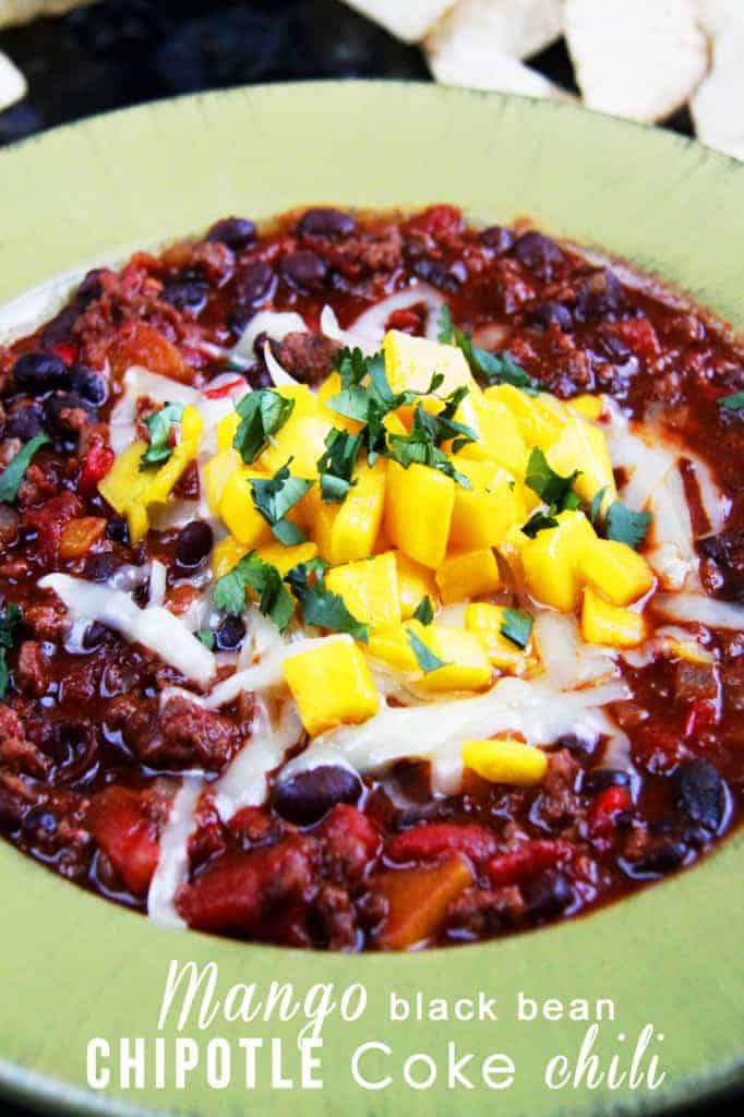A bowl of popular chili topped with diced mango, shredded cheese, black beans, and chopped herbs, served in a green dish. This must try chili is a delicious twist on classic chili recipes.