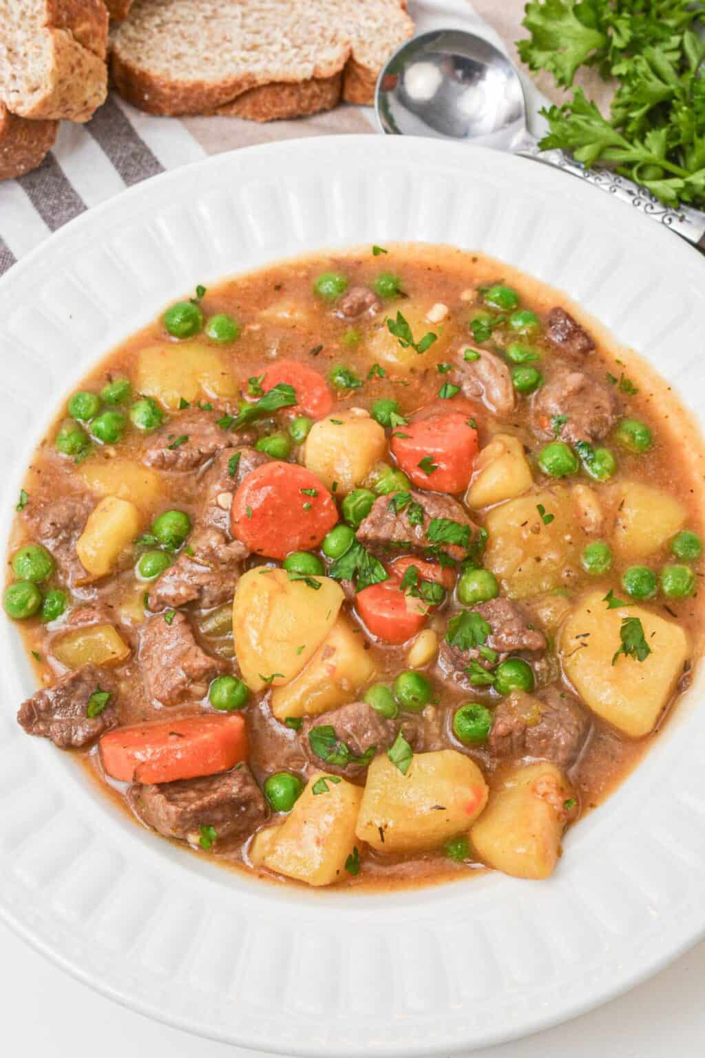 A white bowl filled with Dutch Oven Beef Stew featuring chunks of beef, potatoes, carrots, and green peas, garnished with chopped herbs. Slices of bread and parsley are in the background.